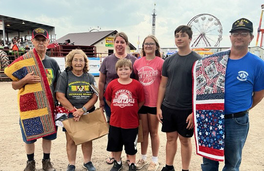 “Quilt of Valor Families 2025”: Veterans Roger Acklie (far left) and Dan Morris (far right) were joined by their families to receive a Quilt of Valor July 10 during the Madison County Fair and Rodeo.